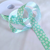 Green floral satin ribbon with a daisy flower print unrolled beside its spool, perfect for crafts and party decorations